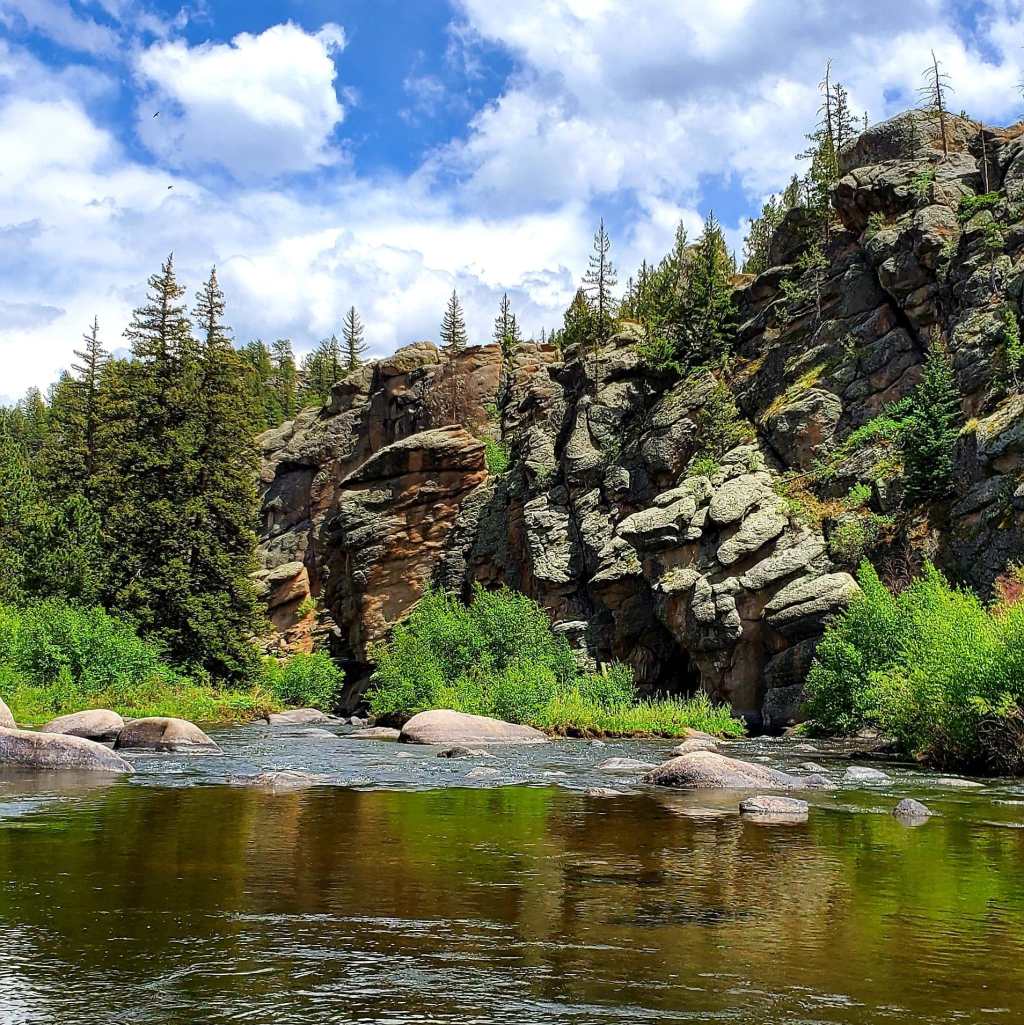 A scenic view of a river surrounded by lush greenery and rocky cliffs under a blue sky with scattered clouds.