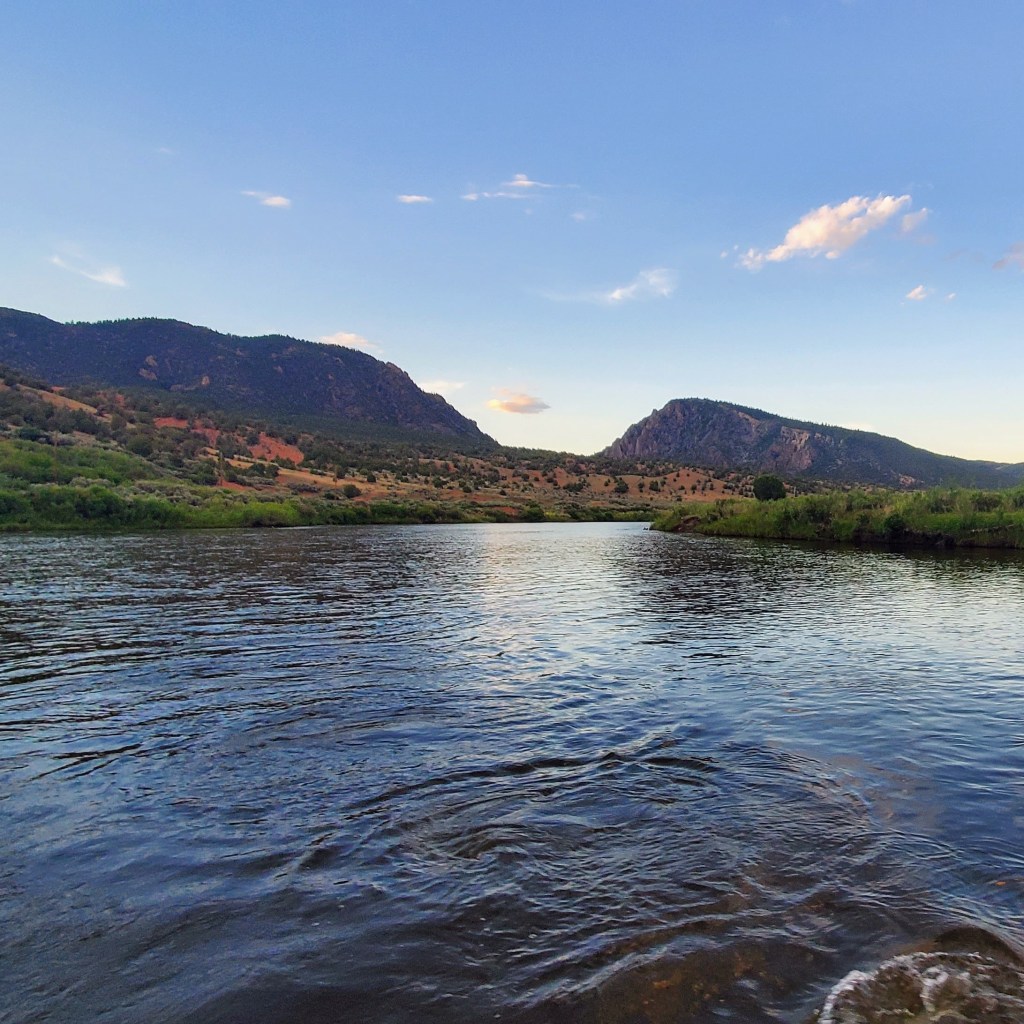 A tranquil river scene surrounded by mountains and greenery under a blue sky.
