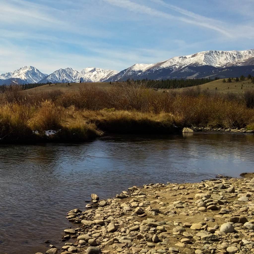 A serene riverbank scene featuring smooth stones and a clear river, with snow-capped mountains in the background and a blue sky overhead.