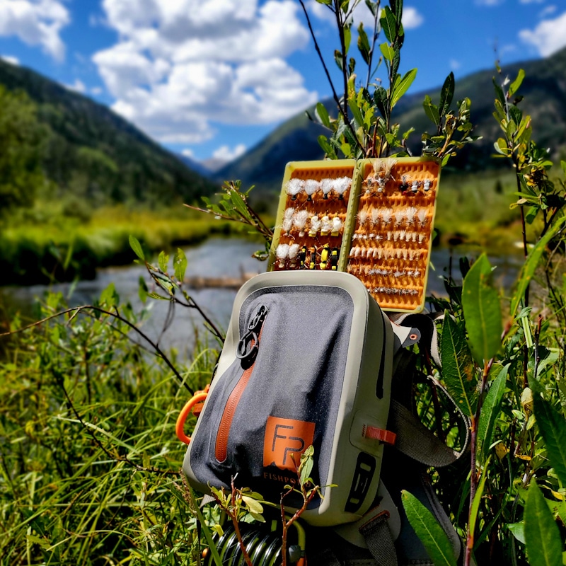 A fishing backpack with a fly box open, displaying various fishing flies, set against a scenic river and mountain backdrop under a blue sky with clouds.