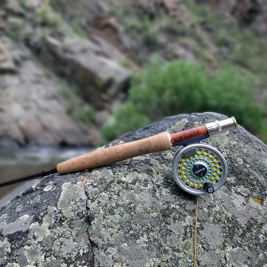 A fly fishing rod and reel placed on a rock near a waterway, surrounded by greenery and rocky terrain.