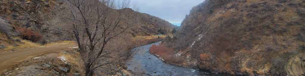 Scenic view of Waterton Canyon featuring a river surrounded by rocky hills and bare trees, with a dirt road running alongside.