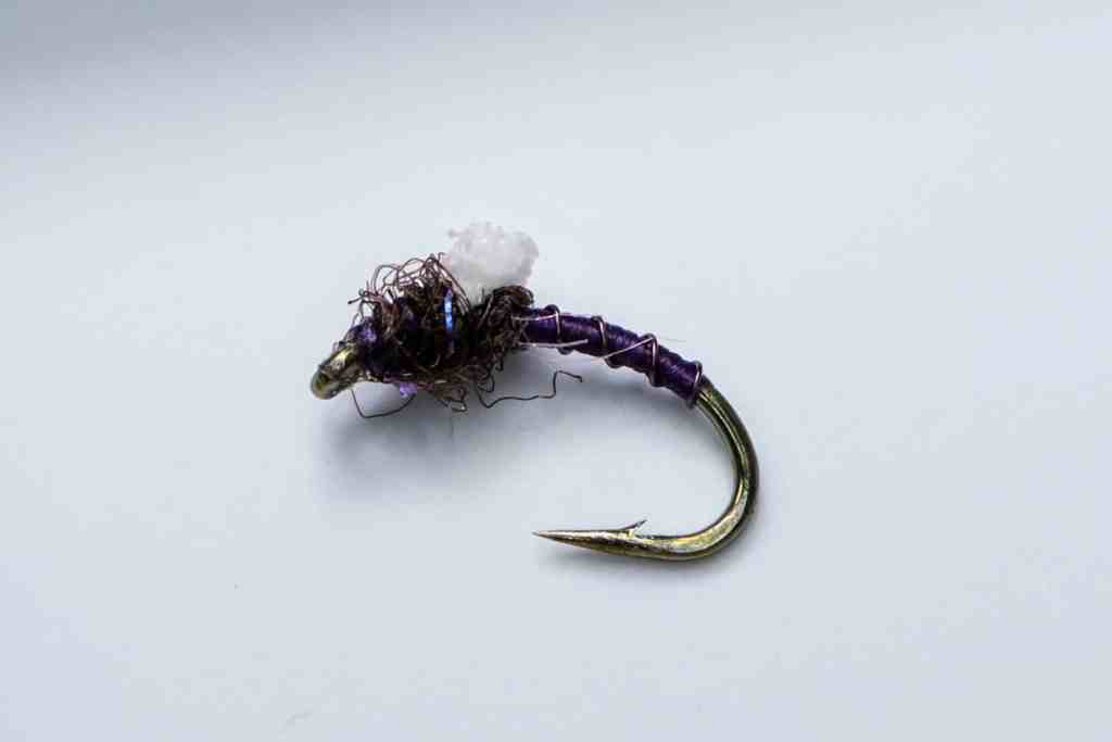 A close-up of a "Foamed Wing Emerger in Purple" fly pattern, tied by Brady Laehr. The fly features a gold hook with a purple wrapped body, a shimmery dark thorax, and a white, foamy wing case at the top. This fly is for sale at Fliesontheline.com.