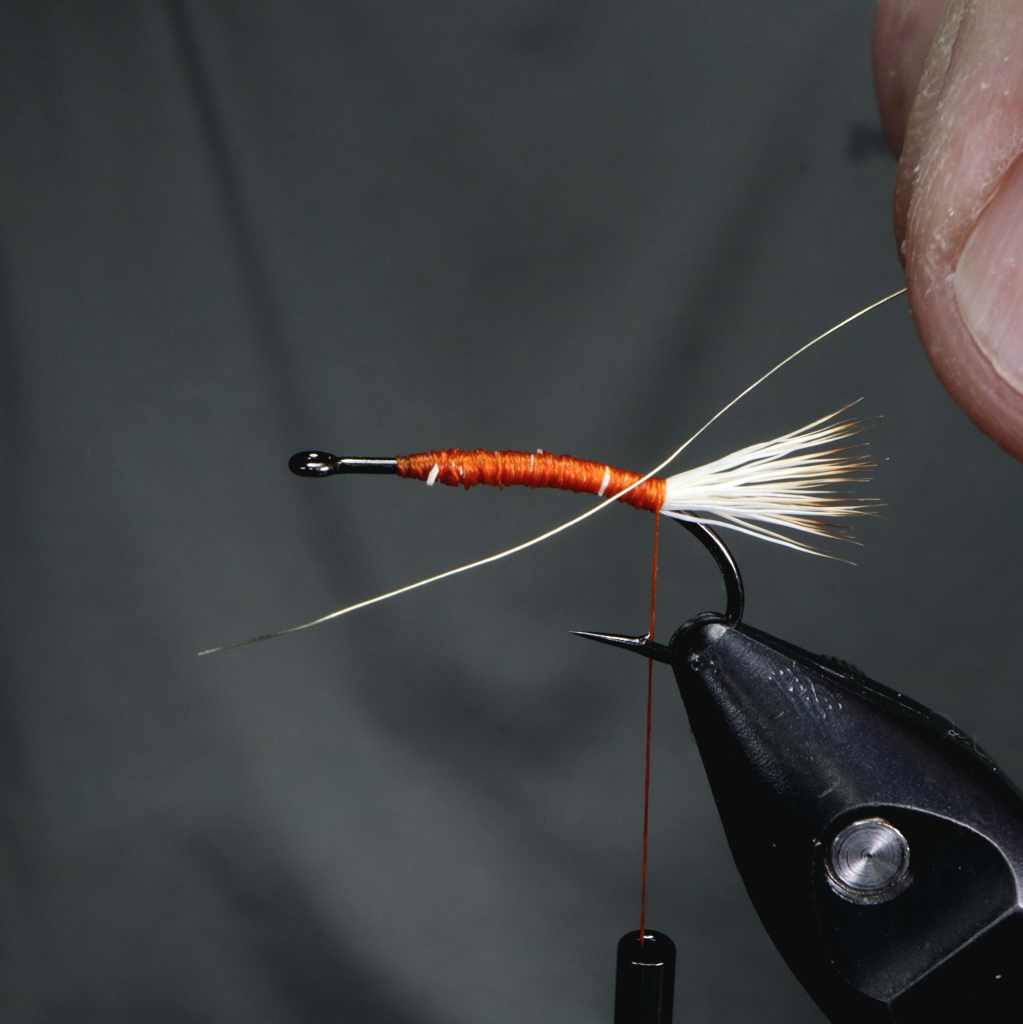 Close-up of a fly tying setup featuring an orange thread base on a hook, with a white elk hair tail positioned above.