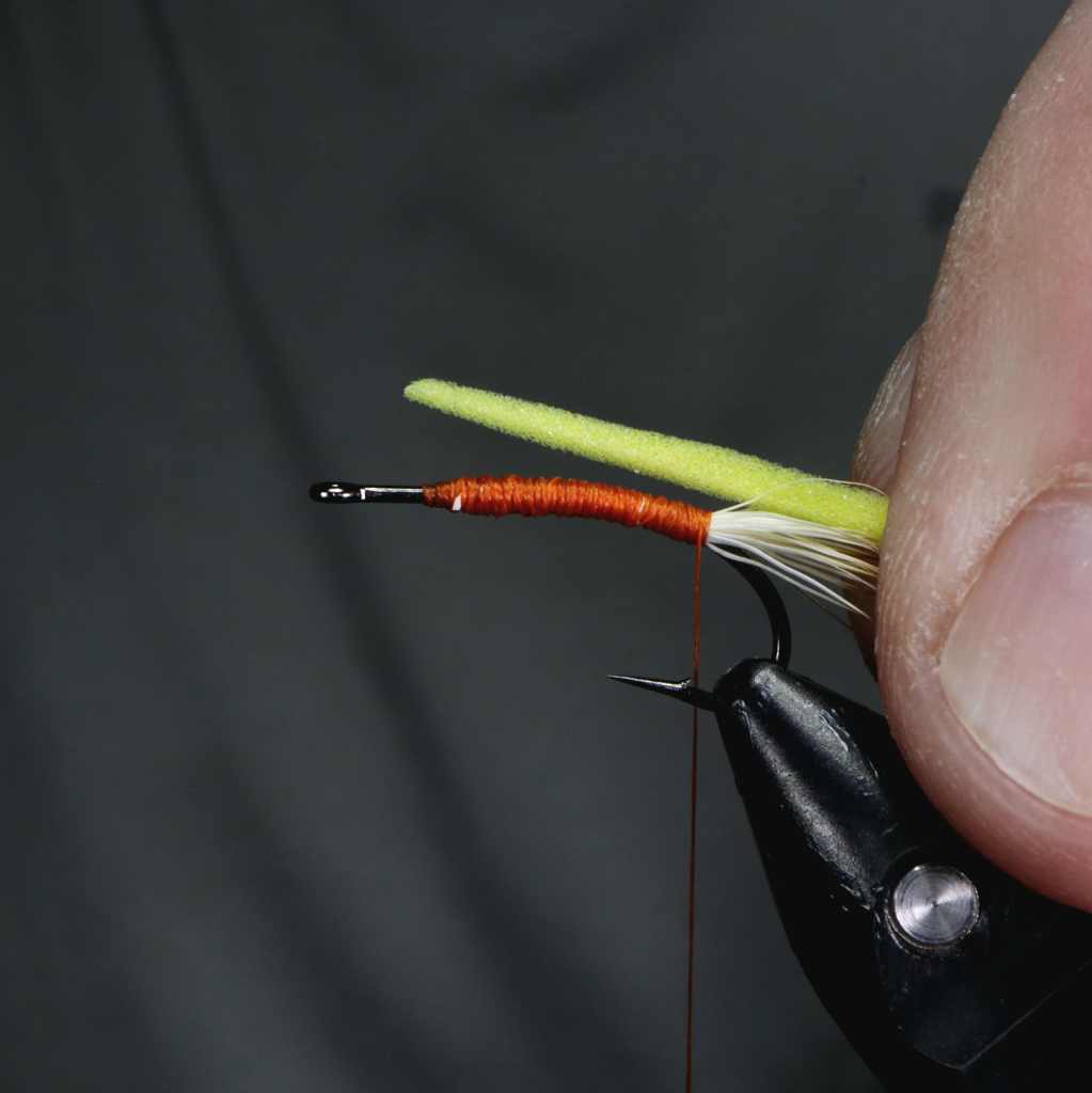 A close-up of a person's hand holding a fishing hook with orange thread wrapped around it, and a green foam piece positioned above. The background is dark, focusing on the fly-tying process.