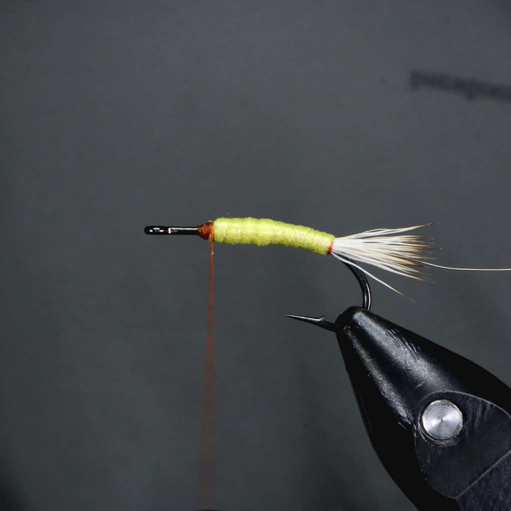 Close-up of a tied fishing fly with a green foam body and a white elk hair tail, resting on a fly tying vise.