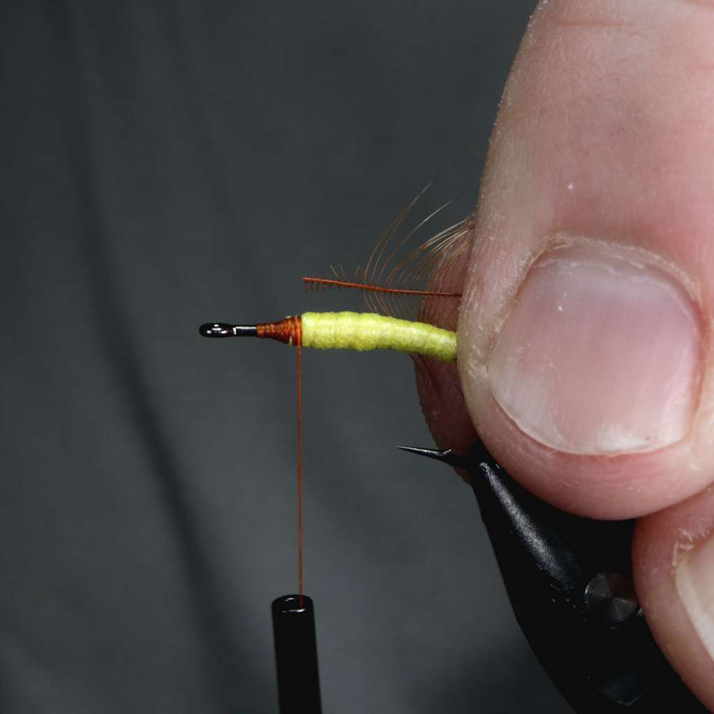 A close-up image showing a person's hand holding a hook with a foam body and thread wraps being secured for tying a fly fishing lure.