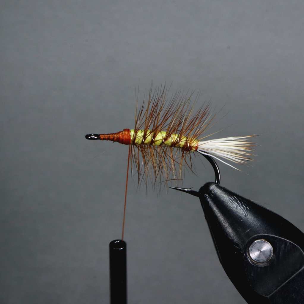 Close-up of a tied dry fly, featuring a foam body in olive and orange, brown hackle, and white elk hair wing, positioned on a fly-tying vise.