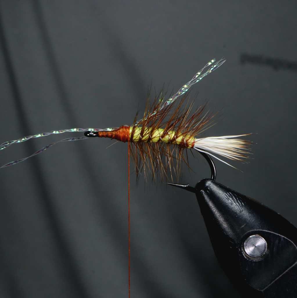 Close-up of a tied dry fly featuring a foam body, with a brown and grizzly hackle, and crystal flash fibers for added effect.