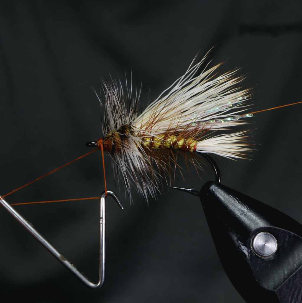 A close-up of a partially tied fly pattern featuring a foam body, bleached elk hair tail and wing, with shiny Krystal Flash fibers. Thread wraps are visible on the hook shank, highlighted against a dark background.