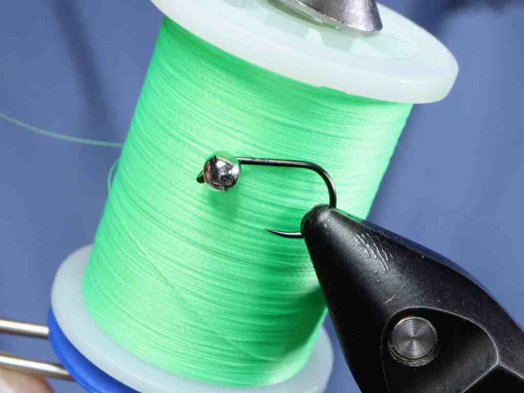 A close-up image of a green spool of thread with a fishing hook attached, held by a pair of pliers.