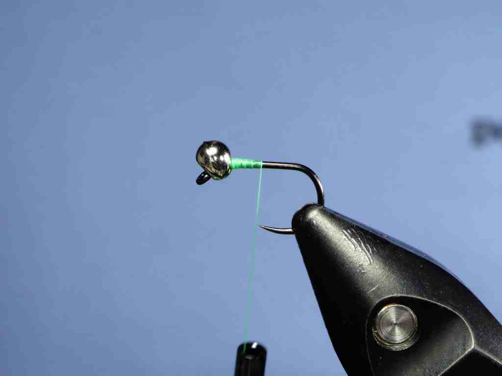 Close-up image of a fishing hook with a shiny metal bead attached, and green thread wound around the hook, set against a light blue background.