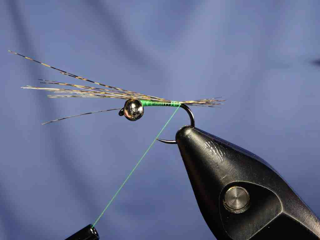 Close-up of a fly tying technique showing a green thread wrapped around a hook with a shiny bead and long tail fibers.