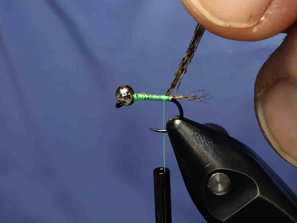 Close-up of a hand tying a fishing fly with a green thread on a hook, featuring a silver bead head and a feather.
