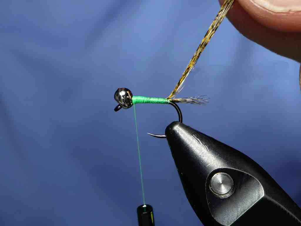 A close-up image of a fishing fly being tied, featuring a green thread wrapped around a hook with a metal bead head, and a person's hand holding a piece of feather.