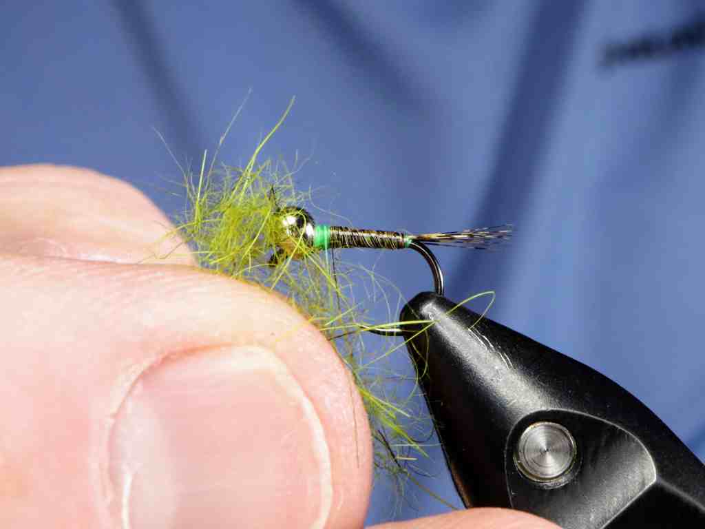 A close-up of a person holding a fly fishing lure with green materials, using a pair of pliers for tying.