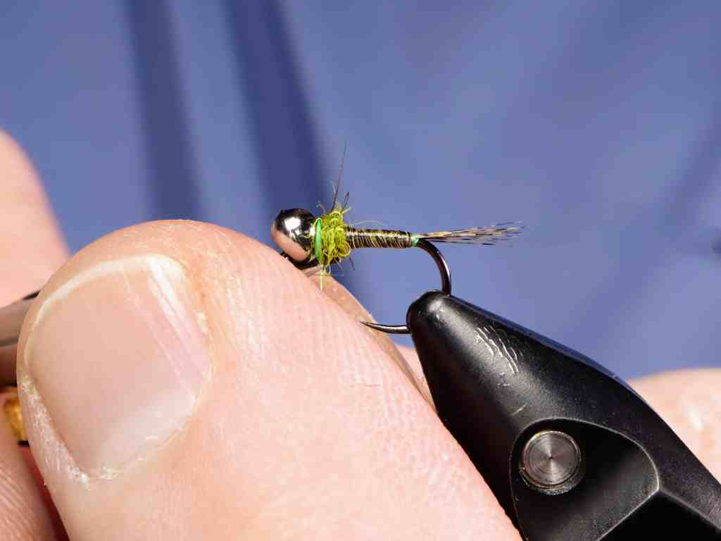 A close-up image of a person tying a fishing fly, showcasing a small green and silver fly attached to a hook, with a focus on the fingers holding the fly-tying tool.