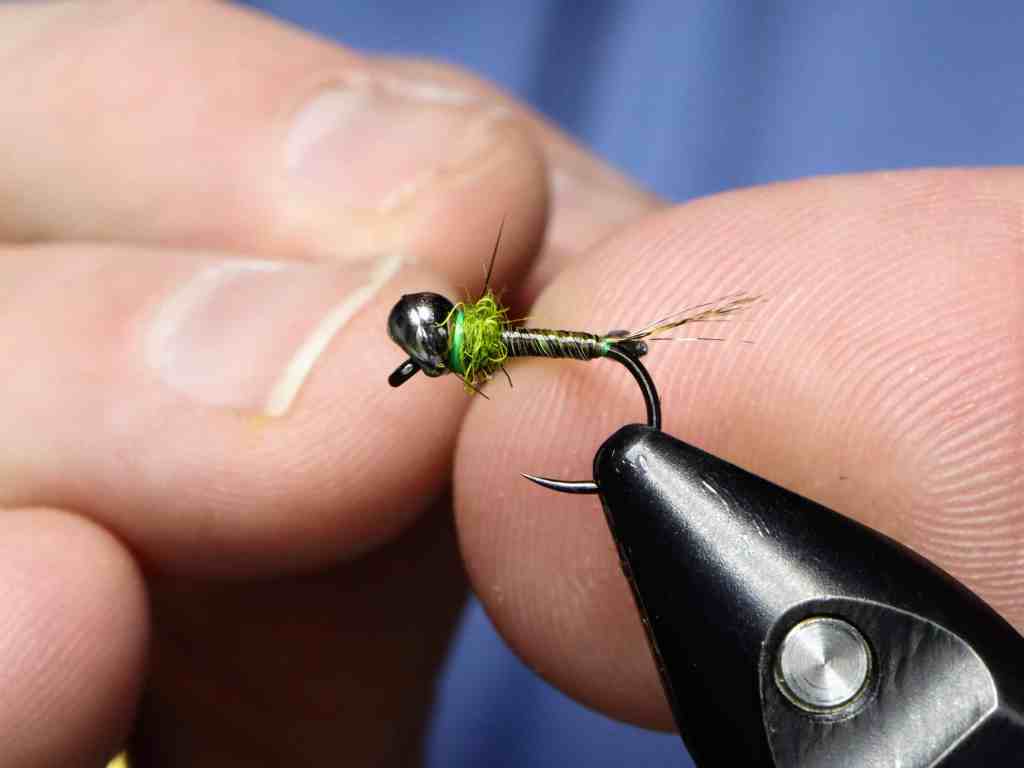Close-up of a person's hand holding a small fishing fly with a green body and black bead, ready for tying or use.