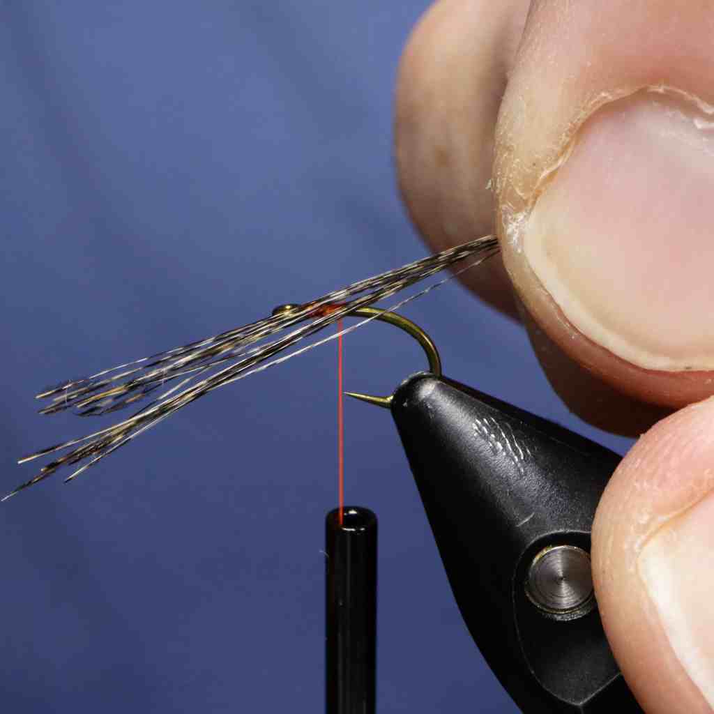 A person holding a pair of scissors, preparing to tie materials onto a hook for fly tying, with strands of Coq De Leon fibers and fluorescent thread visible.