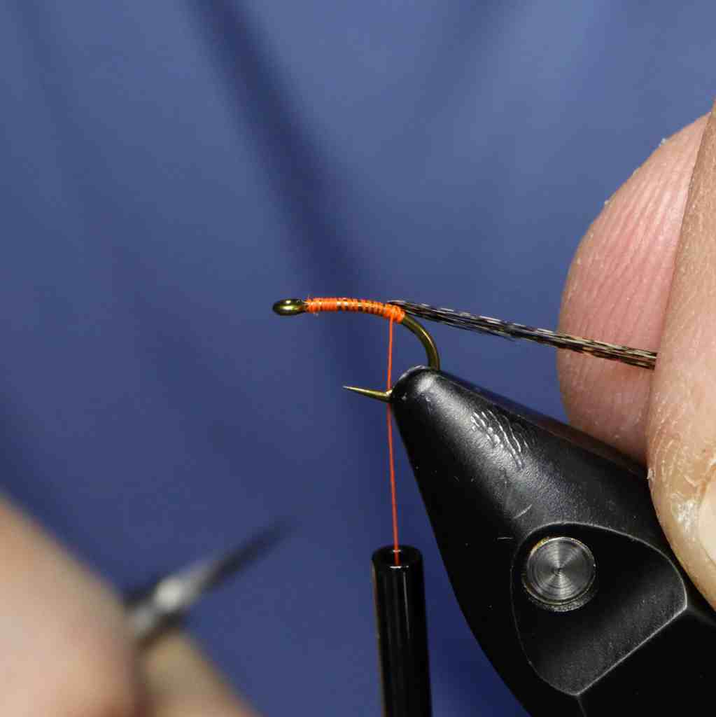 A close-up image showing the process of tying a fly with an orange thread on a hook, held with pliers. The background features a blue surface, emphasizing the focus on the fly tying technique.