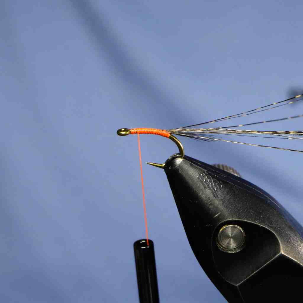Close-up of a fly-tying setup with an orange thread wrapped around the hook shank, preparing to create a fly pattern.