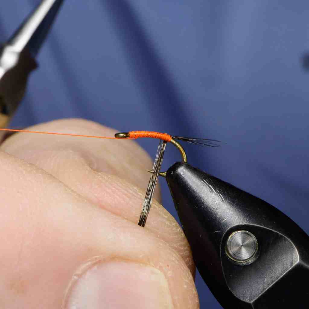 A close-up image showing a person's hand tying a fishing fly with fluorescent orange thread on a hook, illustrating the beginning stages of the fly-tying process.