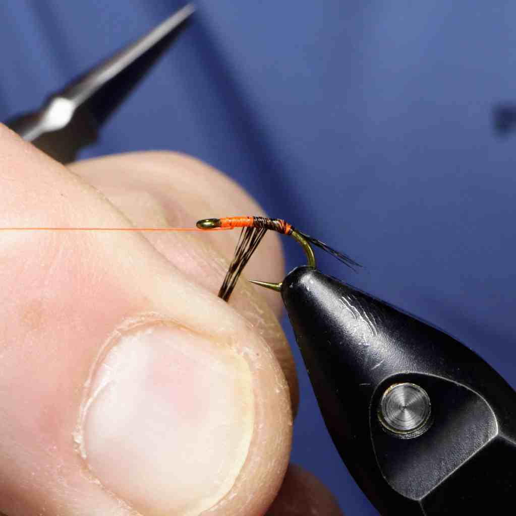 A close-up image of a hand tying a Fl. CDL Nymph fly on a hook using fluorescent orange thread.