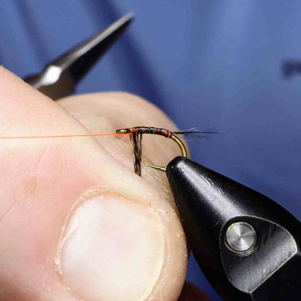 Close-up image of a person's hand tying a fly fishing pattern, showing the thread wrapped around the hook shank with fibers attached.
