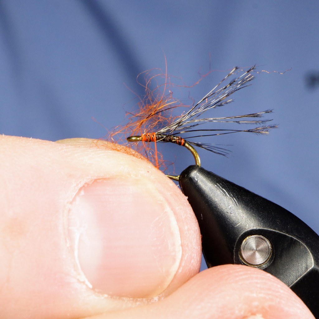 A close-up image of a person tying a fishing fly, showing the hook and thread with some fibers and dubbing at the front.