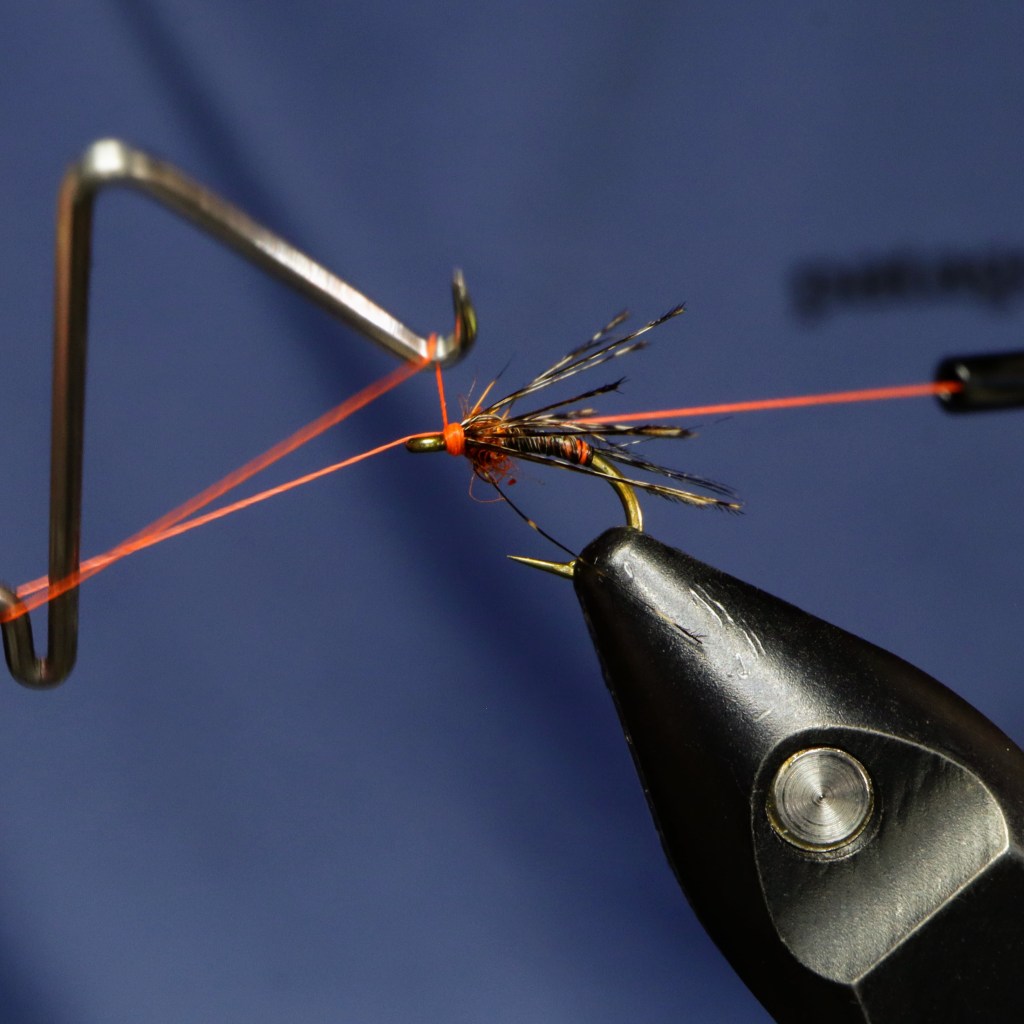 A detailed view of a fly tying process, showing a partially completed Fl. CDL Nymph fly on a hook, with fluorescent orange thread being tied in.