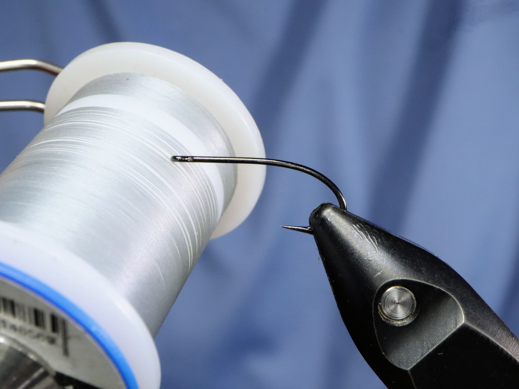 A close-up image of a fishing hook held by a vise next to a spool of fluorescent thread, showing the setup for tying a fly.