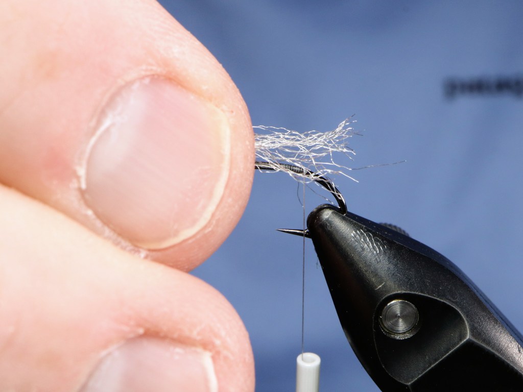 Close-up view of a person's hand holding a fishing hook with thread and feathers being attached, demonstrating the initial steps of fly tying.