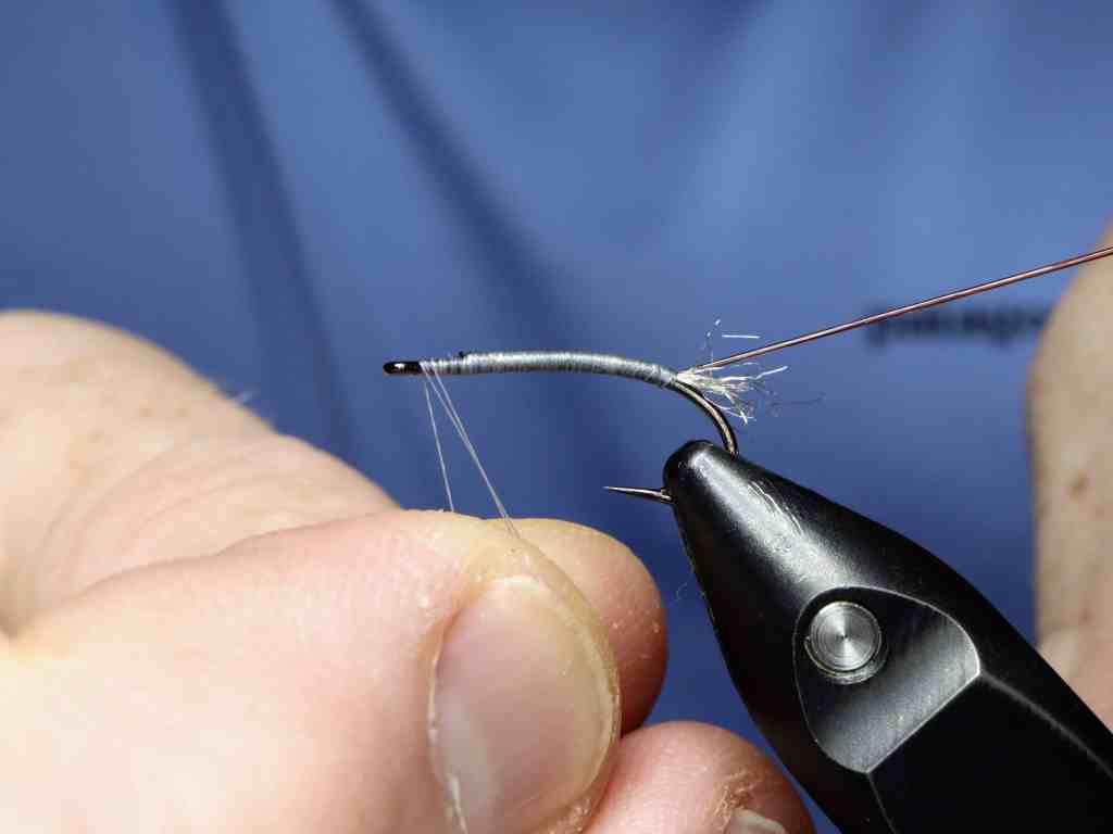 A close-up of a person's hand holding a fly tying vise with a hook secured in place, showing thread being wrapped around the hook.