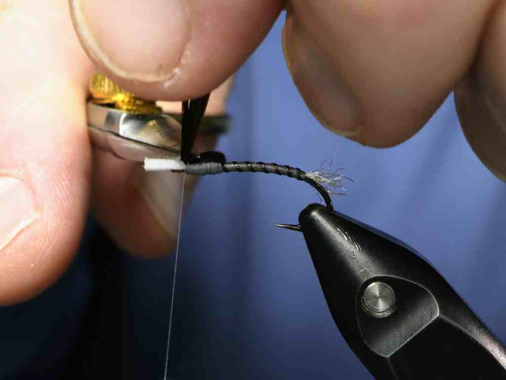 A close-up image of a person tying a fly on a vise, showing a black ribbed body with a pheasant tail thorax and foam extending from the hook.